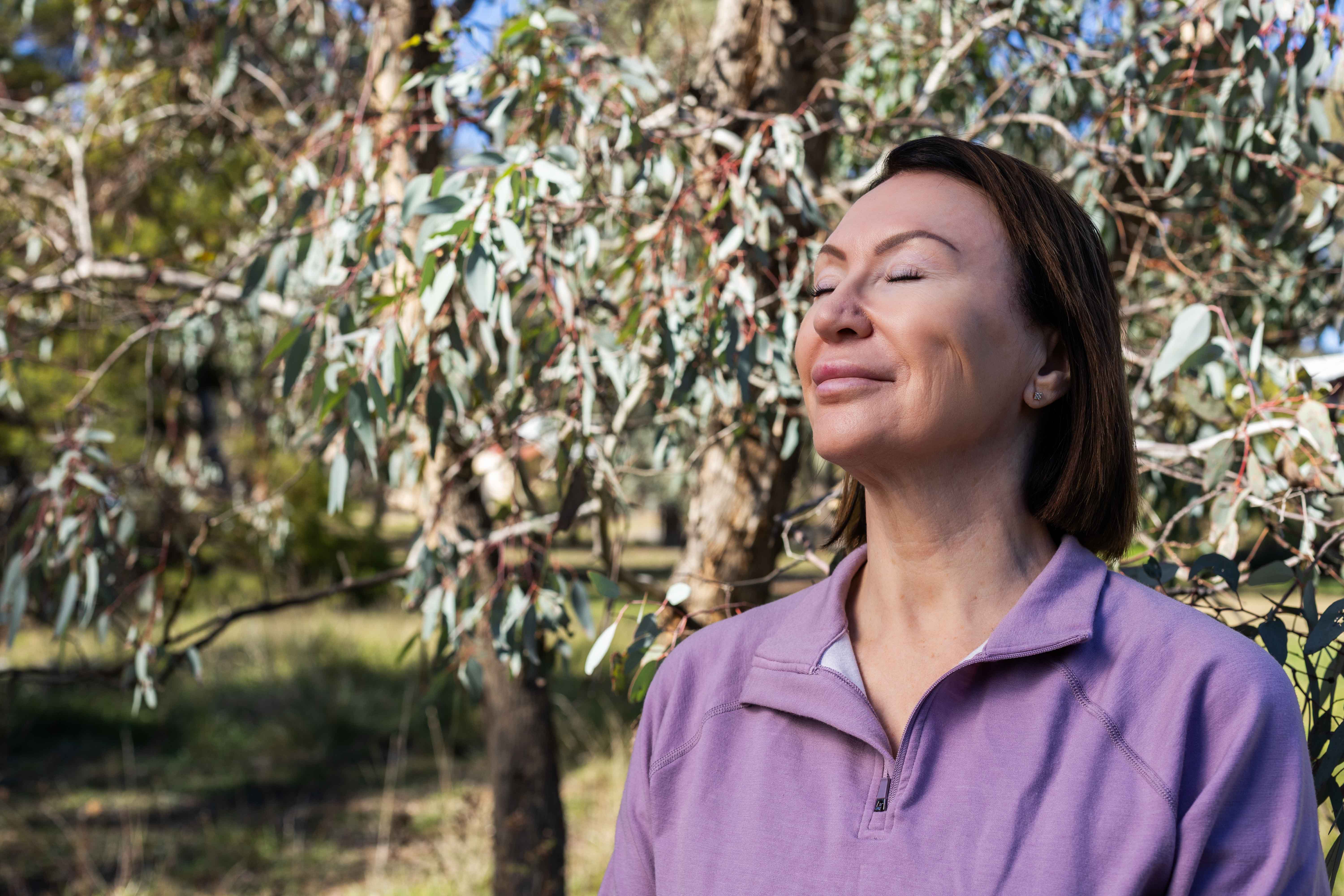 A person standing outdoors among trees with eyes closed, facing the sunlight.
