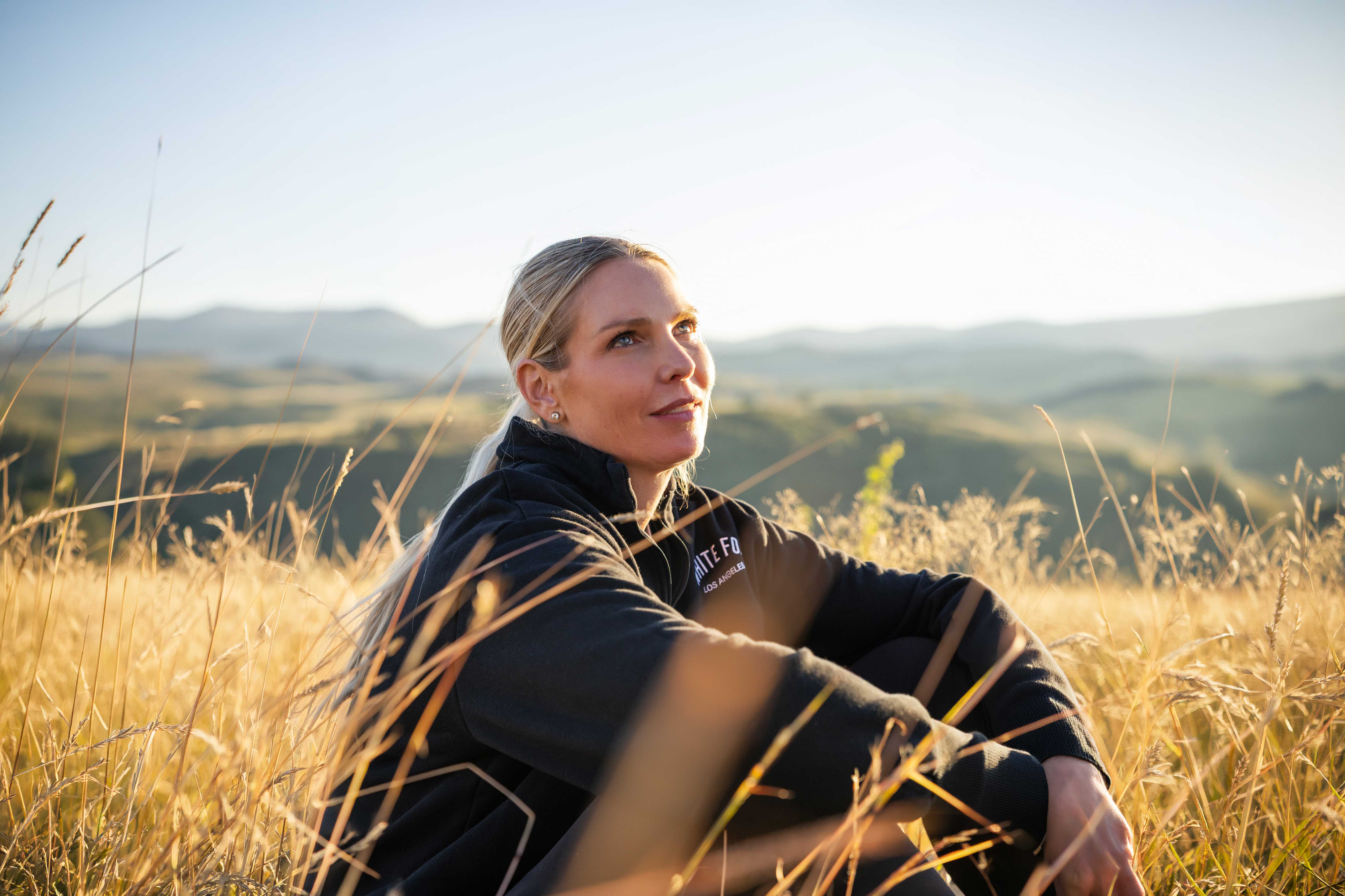Close-up of a person sitting in tall grass on a hillside, looking upward towards the sky.