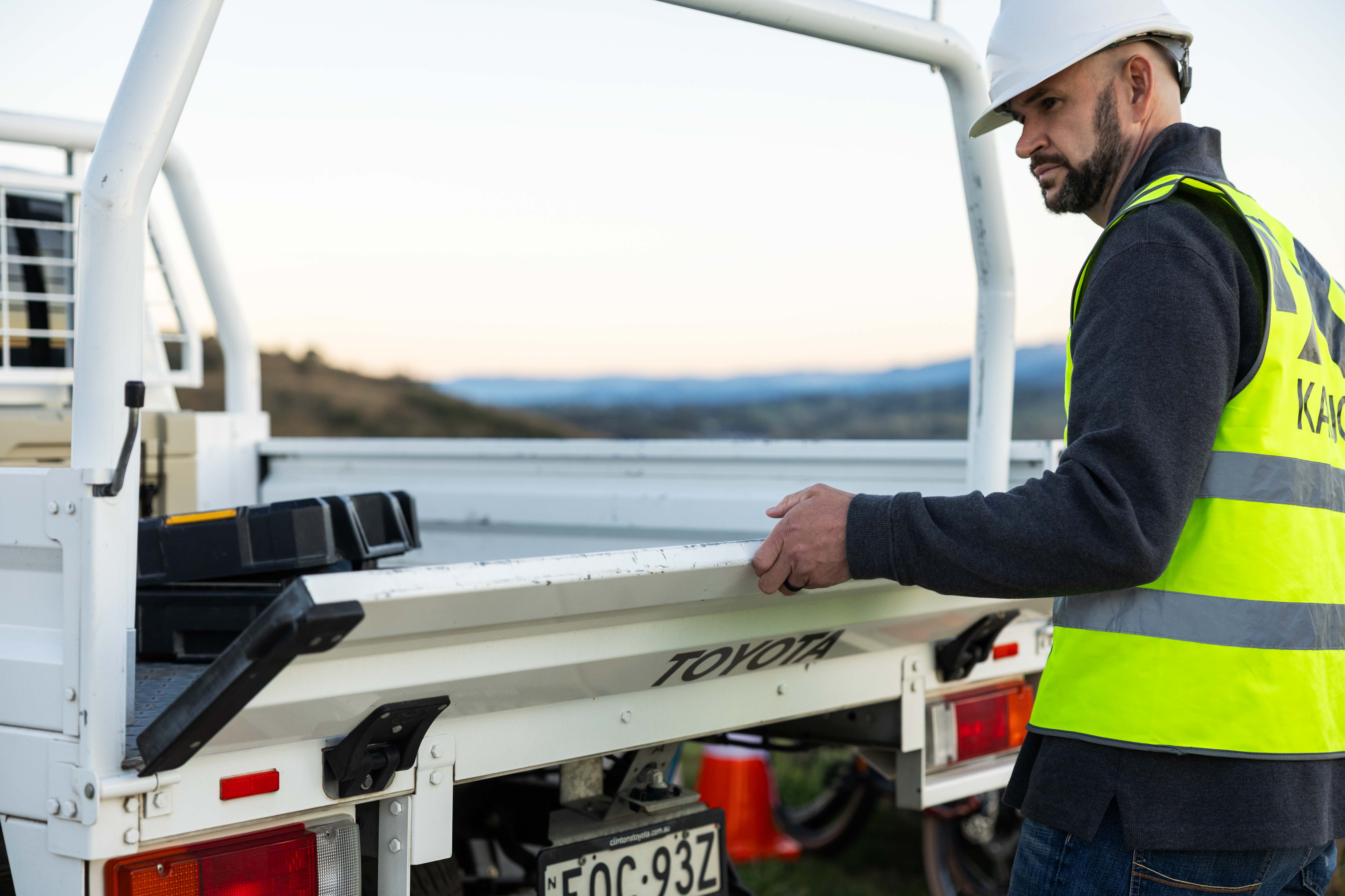 A person wearing a hard hat and high-visibility vest standing at the back of a work vehicle, holding the tray.