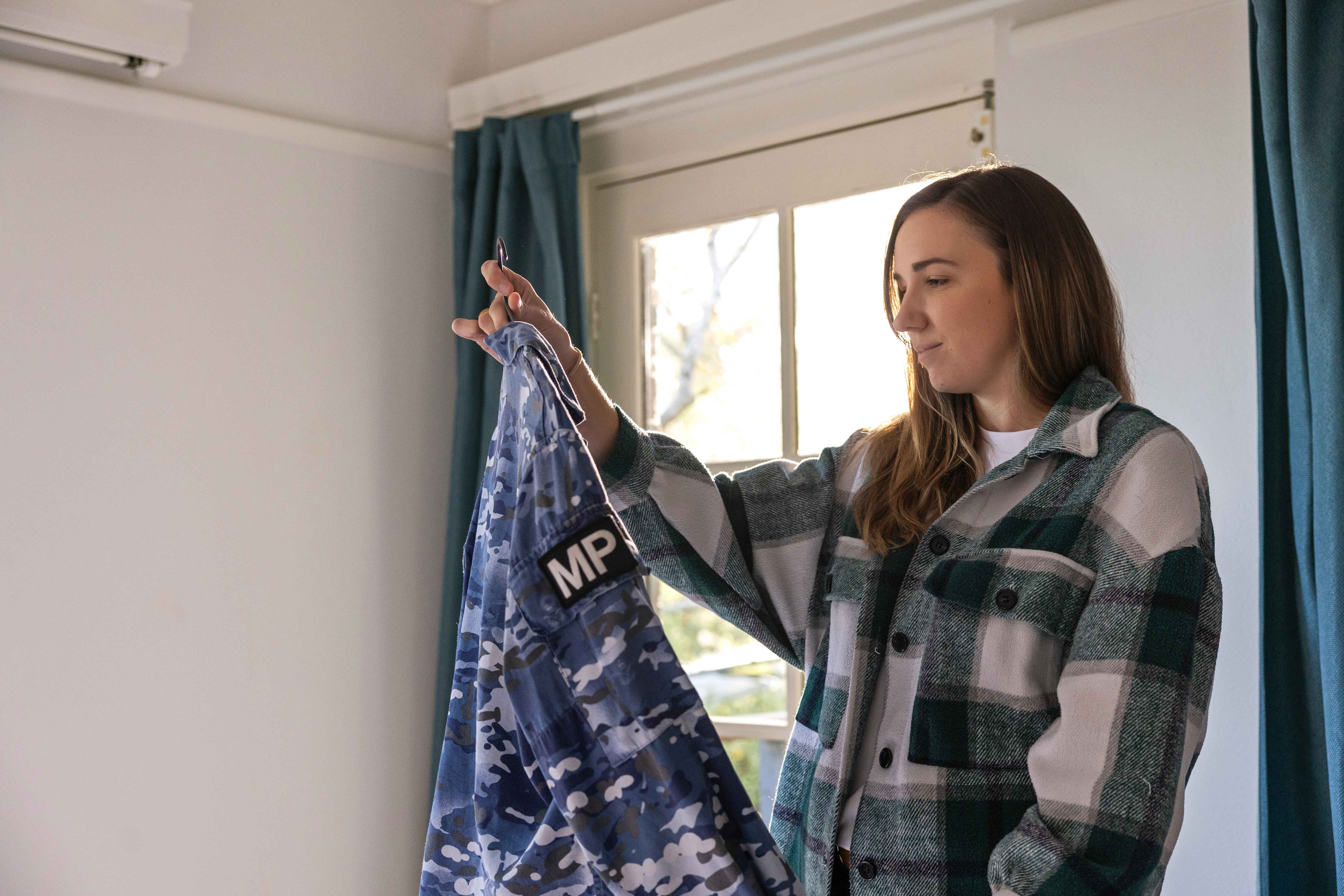 A person holding and looking at their Australian Defence Force uniform on a hanger by a window.
