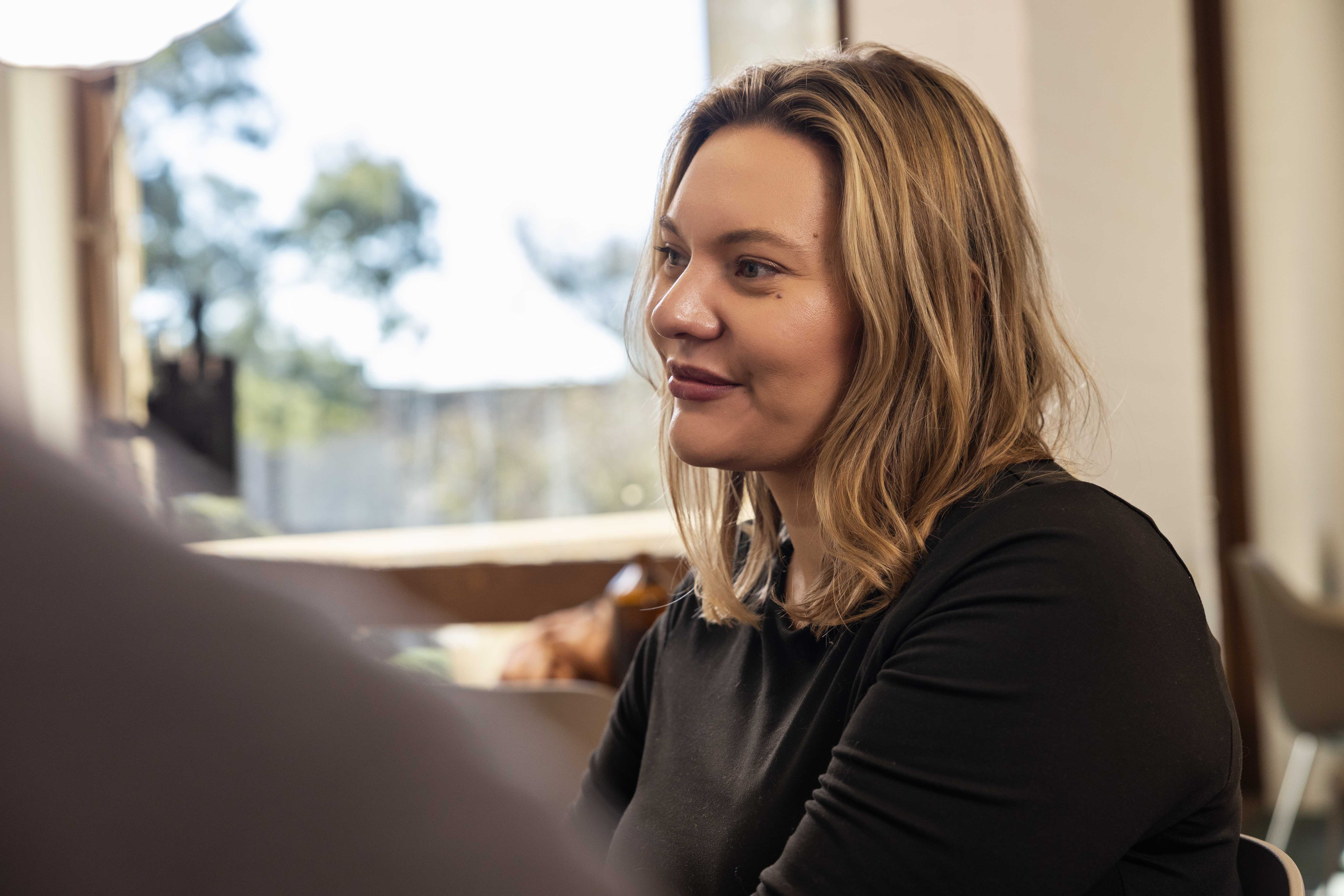 A person sitting indoors, facing and listening to someone in conversation.