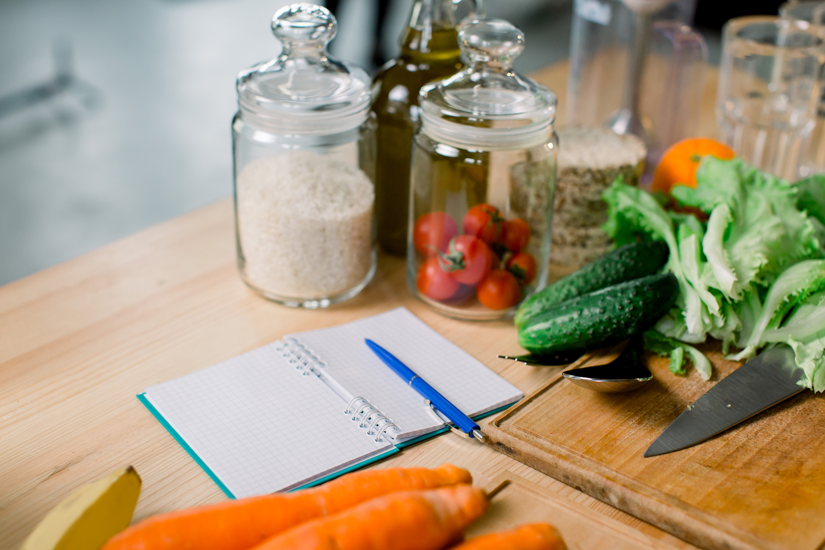 Fresh vegetables, pantry jars and a notebook with a pen arranged on a kitchen bench beside a chopping board and knife.