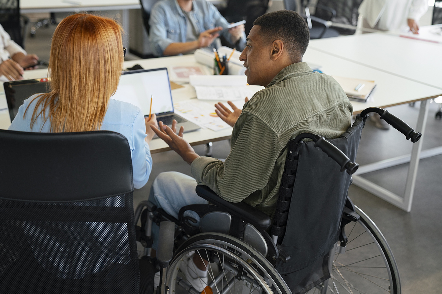 A person in a wheelchair sits at a desk in an office, engaged in conversation with other people around them.