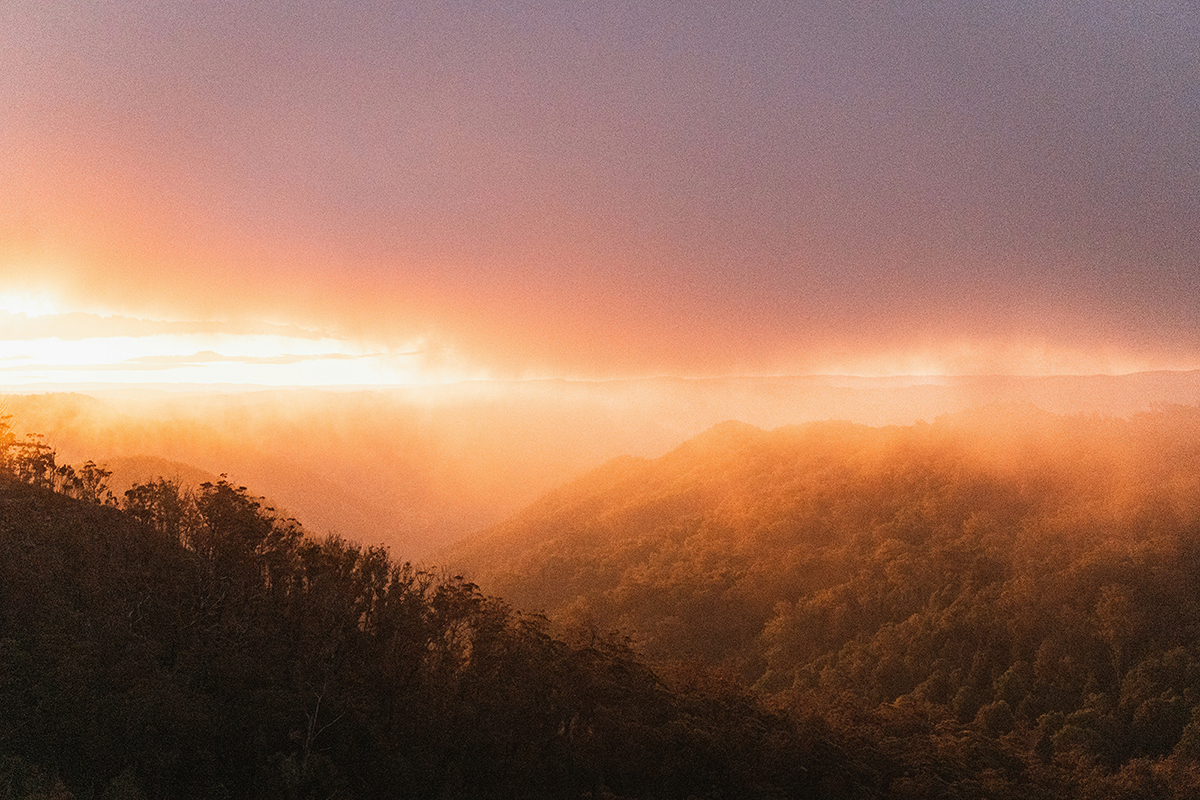 Warm sunset light over misty hills and bushland, with layers of trees and landscape fading into the distance.