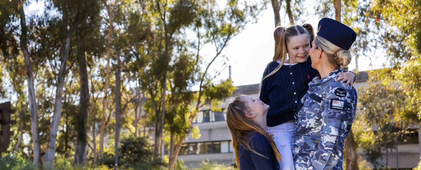 A person in Australian Defence Force uniform holding one child, with another child standing beside them in a park.