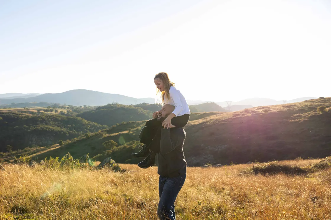 An adult carrying a child on their shoulders while walking through a grassy hillside.