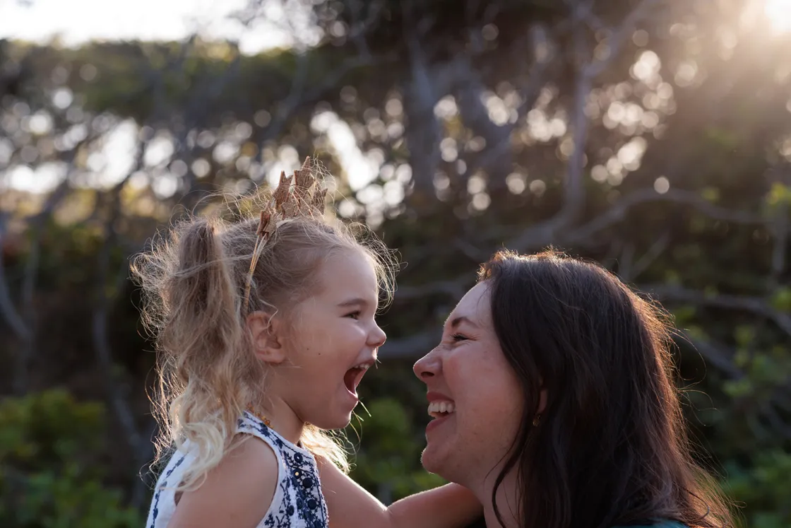 A person lifting and holding a young female child close while facing each other outdoors, with trees and soft sunlight in the background.