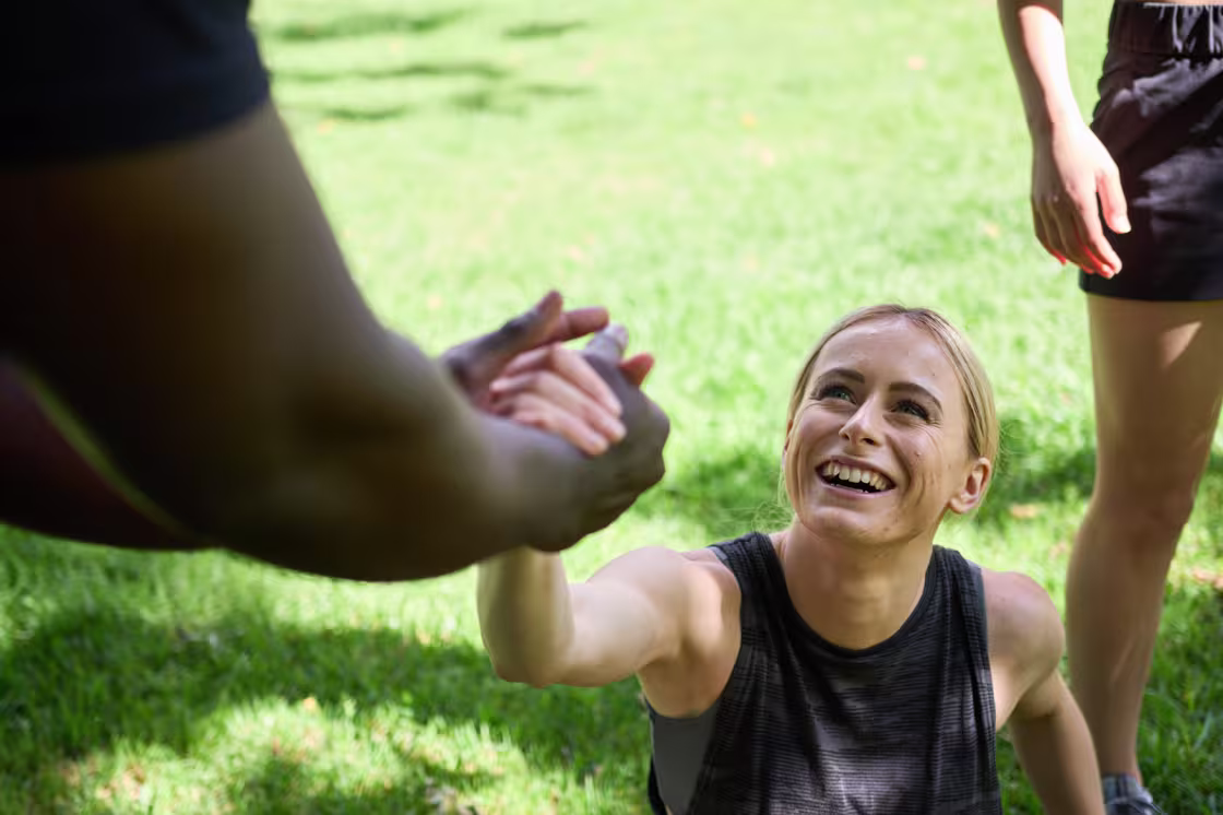 A person sitting on grass during outdoor exercise reaching up to take a helping hand from another participant.