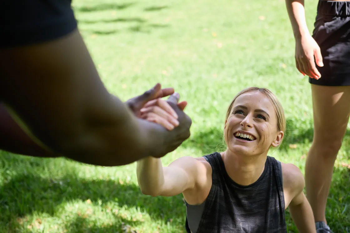 A person sitting on grass during outdoor exercise reaching up to take a helping hand from another participant.