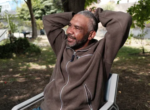 Male sitting on a chair outdoors in a park setting, with arms resting behind their head and trees in the background.