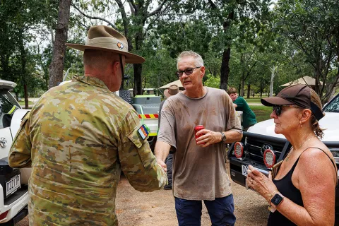 A person in Australian Defence Force  uniform shaking hands with another person outdoors near parked vehicles, with others standing nearby in a bushland setting.