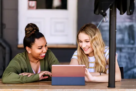 Two adult females sitting together at a table using a tablet in a café or outdoor setting.