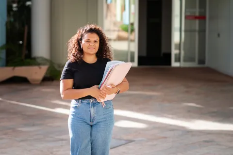 Adult female standing outside a building holding a folder, with doors and a walkway behind.