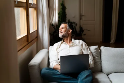 A person sitting on a sofa indoors with a laptop, looking towards a window with natural light.