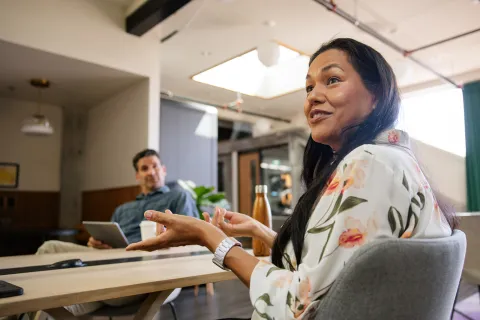 Person seated at a table indoors speaking and gesturing, with another person in the background holding a tablet.