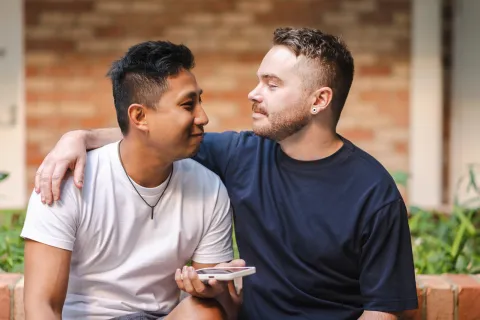 Two people sitting close together outdoors, with one person resting an arm on the other’s shoulder while facing each other.