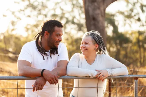 Two people leaning on a fence outdoors and facing each other, with trees and warm sunlight in the background.