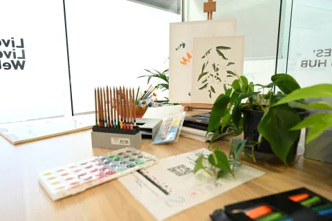 Art supplies arranged on a desk near a window, including coloured pencils, paint and brushes, with a plant and artwork on a stand at the Caboolture Veteran and Family Hub.