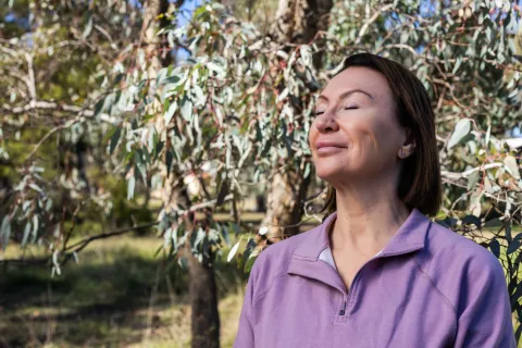 A person standing outdoors among trees with eyes closed, facing the sunlight.
