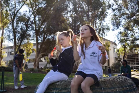 Two young children sitting outdoors in a park, blowing bubbles.