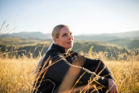 Close-up of a person sitting in tall grass on a hillside, looking upward towards the sky.