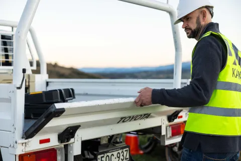A person wearing a hard hat and high-visibility vest standing at the back of a work vehicle, holding the tray.