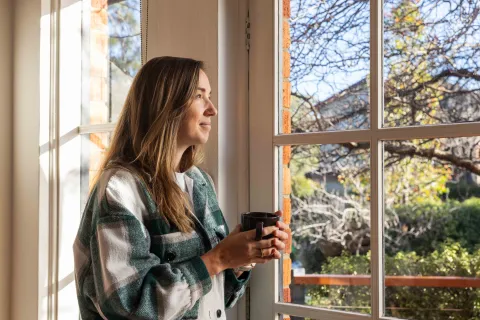 A person standing by a window holding a mug and looking outside.