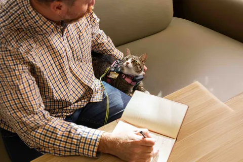 A person writing in a notebook at a table, with an assistance cat beside them.