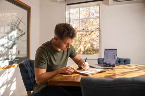 A person sitting at a table writing in a notebook, with a laptop nearby.