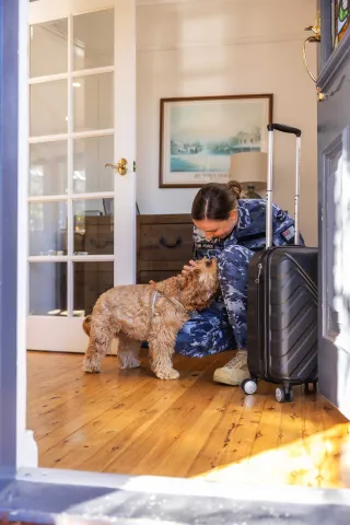 A person in an Australian Defence Force uniform kneeling to greet a dog beside a suitcase at home.
