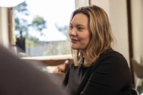 A person sitting indoors, facing and listening to someone in conversation.