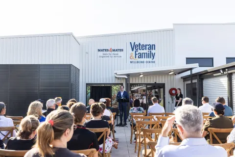 Community gathering outdoors at the Veteran and Family Wellbeing Centre, with a speaker addressing an audience seated in front of the building.