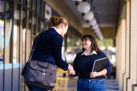 Two people shaking hands in a covered outdoor walkway, with one carrying a shoulder bag and the other holding a folder or notebook.