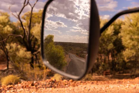 A winding road seen in a vehicle side mirror, with surrounding bushland and trees visible along the roadside under a partly cloudy sky.