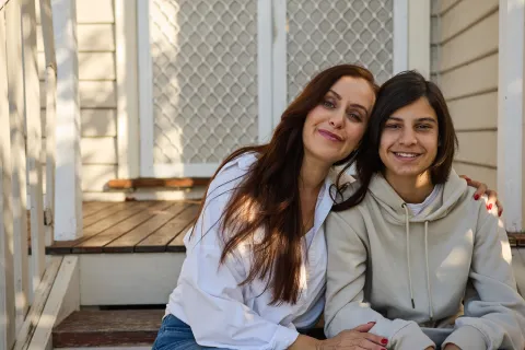 An adult and teenager sitting side by side on front steps, leaning close with an arm around each other.