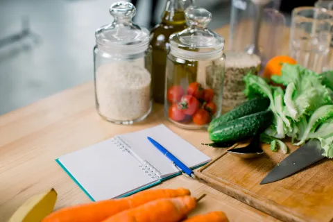 Fresh vegetables, pantry jars and a notebook with a pen arranged on a kitchen bench beside a chopping board and knife.