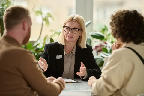 A person in a meeting explaining something to two others seated across a table, with documents in front of them.