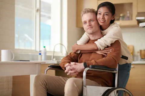 A person in a wheelchair sitting at a kitchen table while another person stands behind with arms around their shoulders.