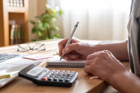 Close-up of a person writing in a notebook with a pen at a desk, with a calculator and laptop nearby.