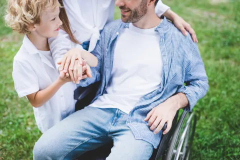Close-up of a child and an adult in a wheelchair holding hands with another person, standing together outdoors.