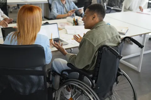 A person in a wheelchair sits at a desk in an office, engaged in conversation with other people around them.