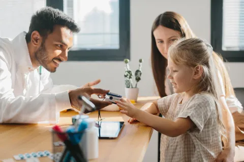 A doctor showing a medical device to a child at a table, with another adult sitting beside them.