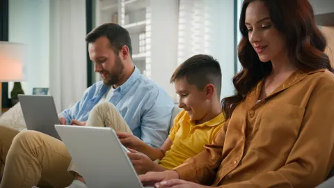 Two adults and a child sitting together on a couch using laptops and a tablet in a home setting.