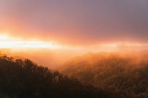 Warm sunset light over misty hills and bushland, with layers of trees and landscape fading into the distance.