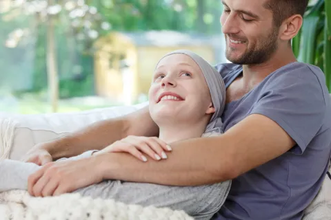 A person sitting on a couch, being hugged from behind by another person resting their arms around their shoulders.