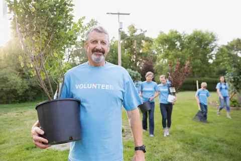 A person carrying a potted tree in a grassy outdoor area, with several volunteers in matching shirts walking behind with plants and tools.