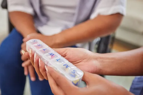 A person holding a labelled weekly medication organiser being passed to another person.