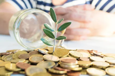Small plant sprouting from a pile of coins, with a glass jar lying on its side and hands blurred in the background.