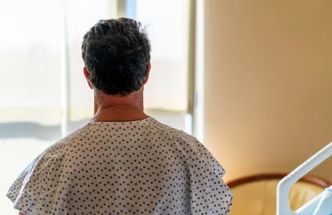 A person wearing a hospital gown sitting on a bed and looking out a window in a hospital room.