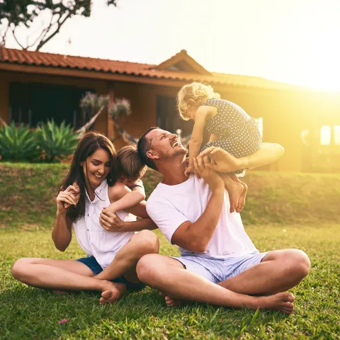 A family sitting on grass outdoors playing together, with one adult lifting a child while others sit nearby in front of a house.